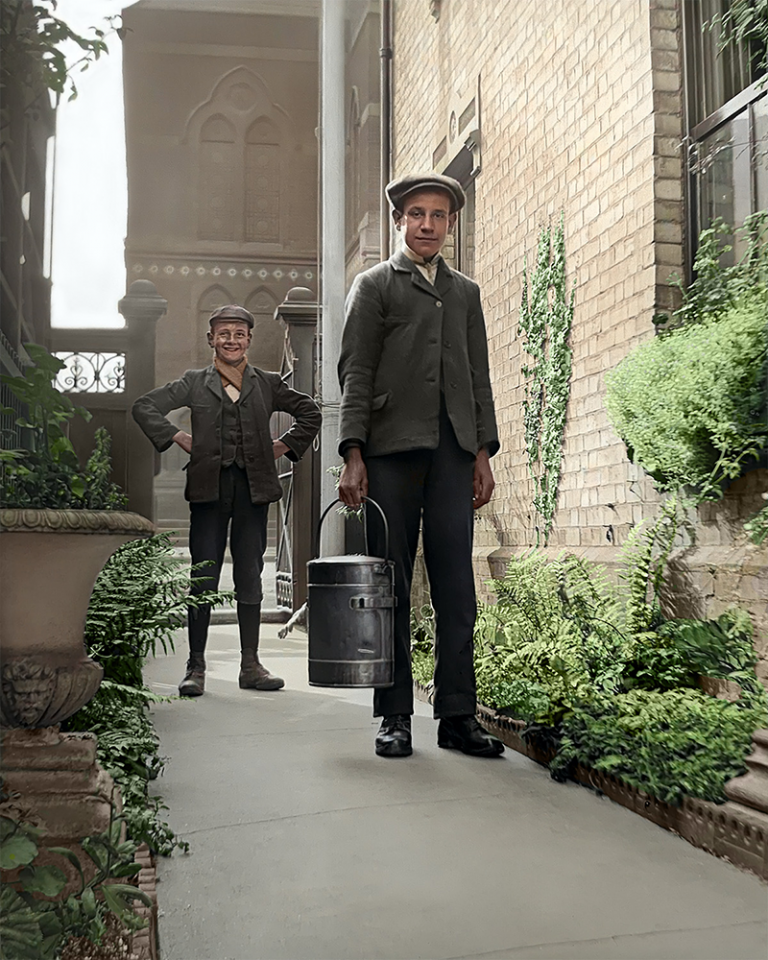 A colourised photo of two Edwardian delivery boys in a passage way of a house with an ornate gate and church in the background. The boy in front carries a milk churn. The boy behind him stands with his hands on his hips and a cheeky smile.