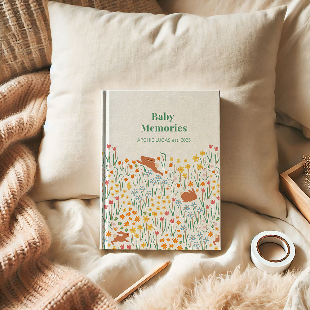 A cream hardback journal with the title, Baby Memories, illustrated with rabbits and flowers sits propped up against a pillow in a cosy bed scene, surrounded by cushions, woollen throw and stationary.