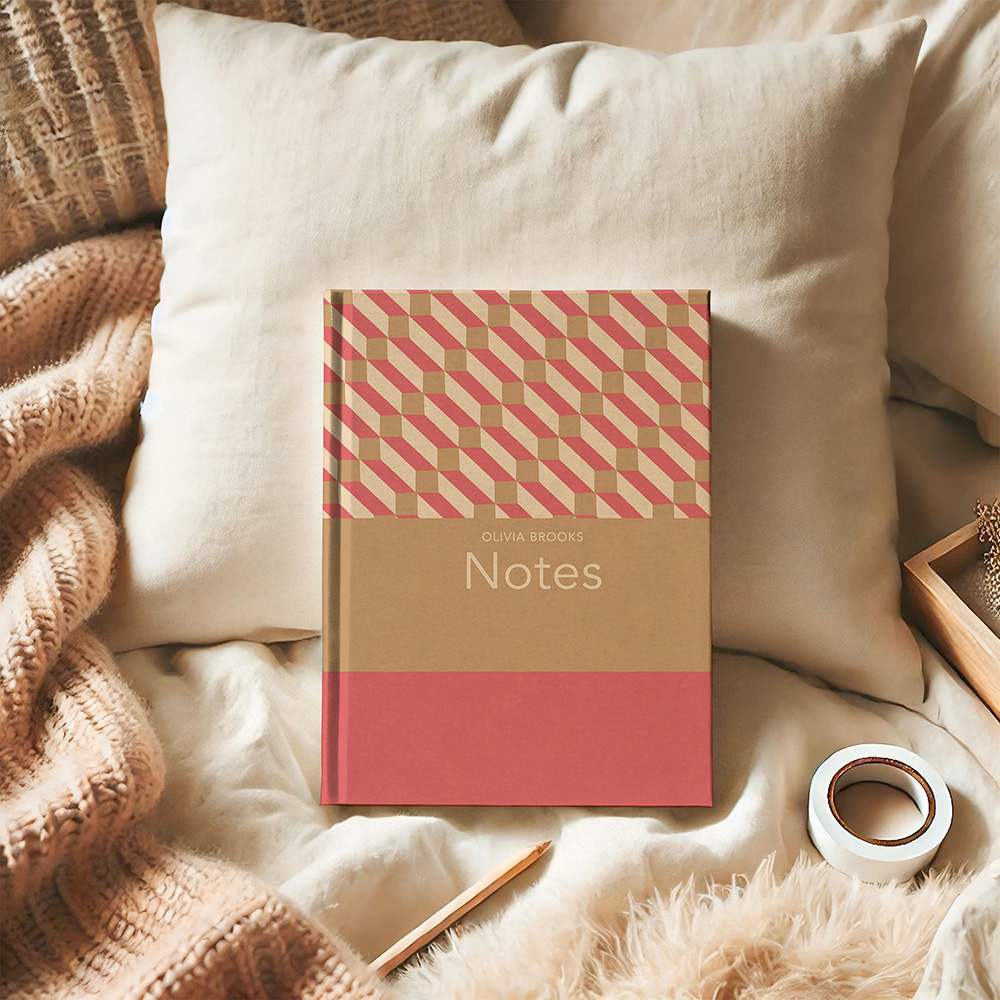 A hardback journal with a cover of bands of brown and coral and a geometric pattern in upper section. The journal sits propped up against a pillow in a cosy bed scene, surrounded by cushions, woollen throw and stationary.