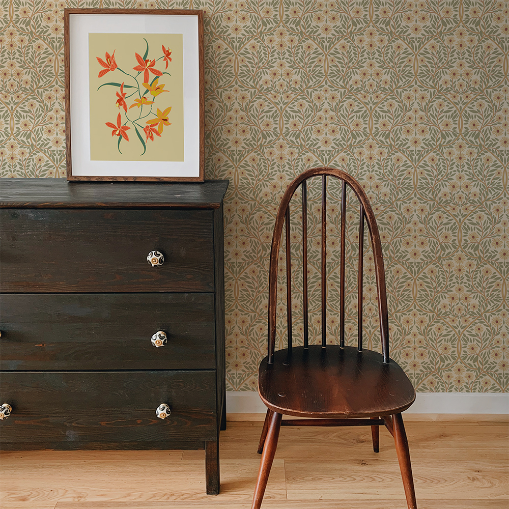 A room with a dark wooden chest of drawers and dark wooden antique chair with a spindle back. On top of the chest of drawers is a framed print of yellow and peach coloured orchids. The wallpaper is an Arts and Crafts style repeat pattern of leaves and flowers called 'Michaelmas Daisy'.
