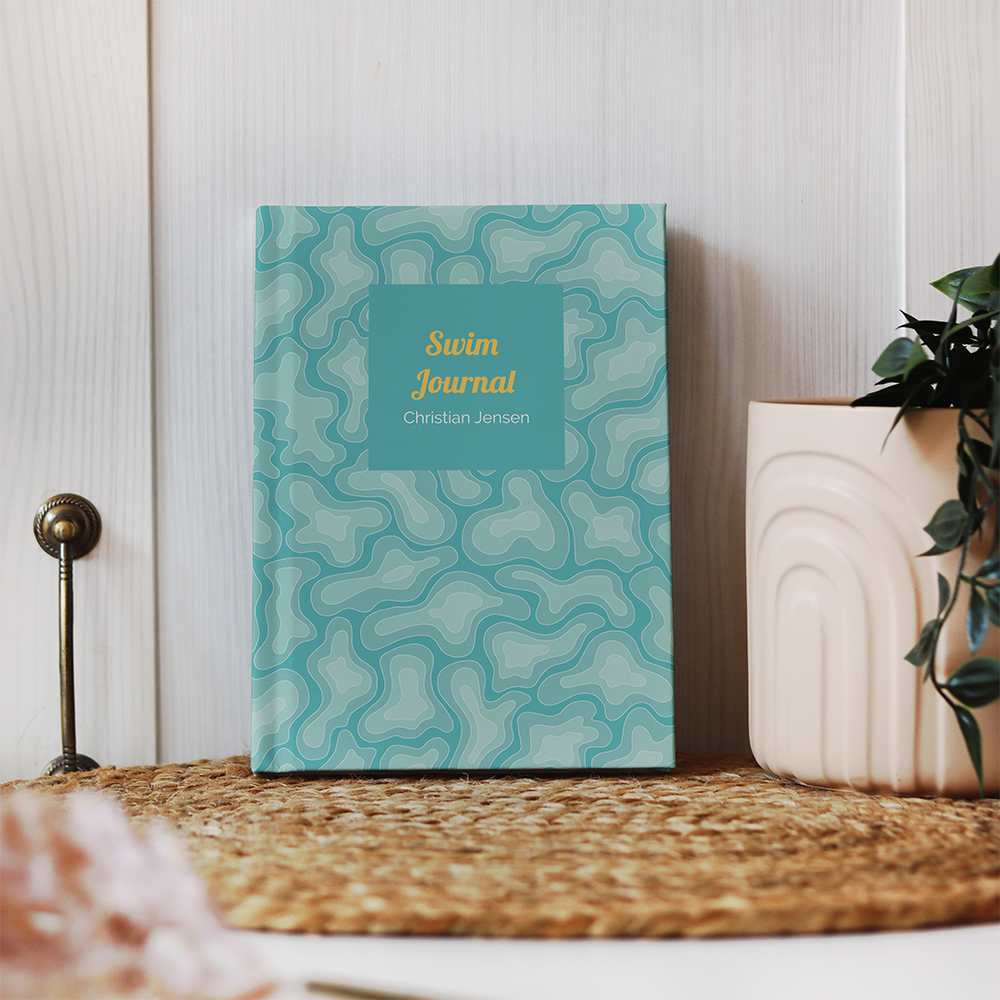 A custom hardback journal with a repeat water camo pattern. In the middle of the design is a blue rectangle with yellow text, 'Swim Journal'. The journal is propped up against the wall on a grass mat next to a potted plant.