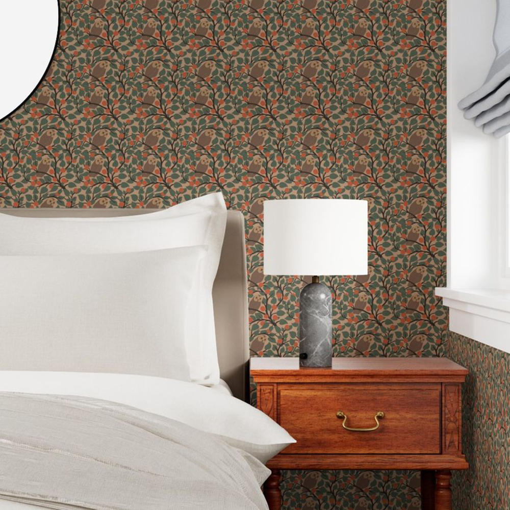 A bedroom scene showing part of a bed with white linen, a bedside table with a small marble lamp with a white lampshade. The wallpaper features a repeat pattern of barn owls sitting in the branches of trees surrounded by fruits of berries