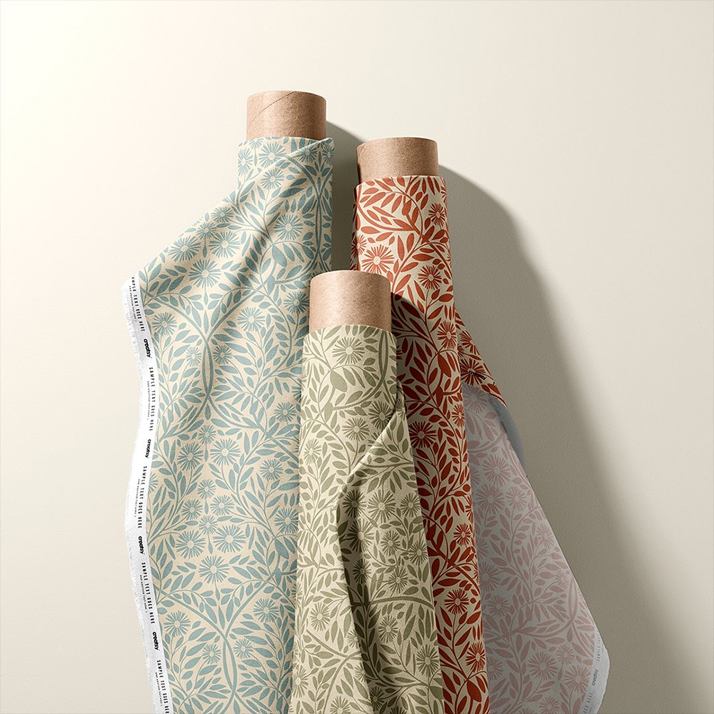 Three upright rolls of fabric on a cream background. The same repeat floral pattern is on each roll but each roll shows a different colourway of the design, 'Michaelmas Daisy'. There is a blue version on the left, in the middle is the fawn version and on the right there is an orange version.