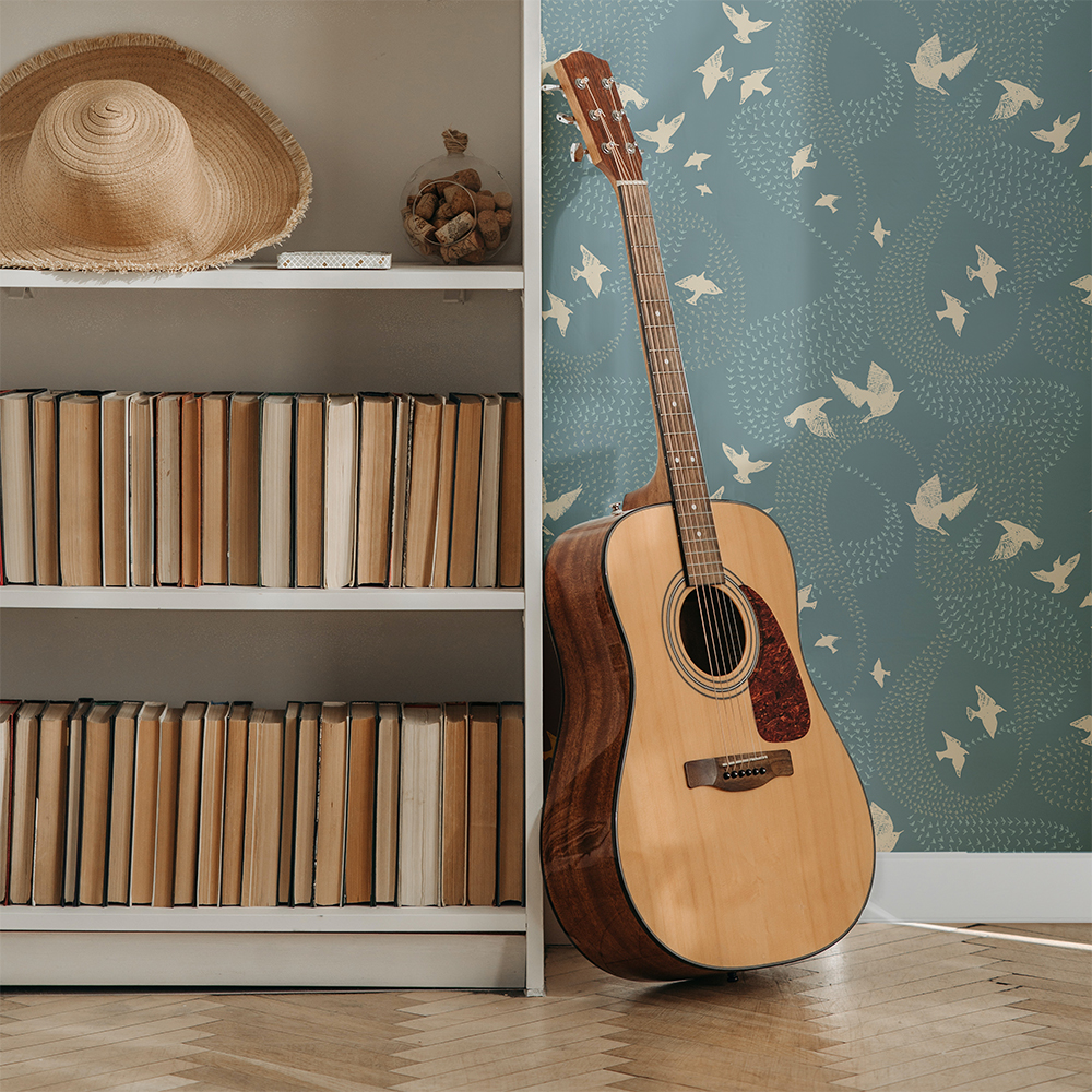A living room scene which shows part of a bookcase with books shelved with the pages facing outside and a hat on one shelf. A guitar leans up against the bookcase The blue wallpaper features murmurations in swirls with close ups of white birds.