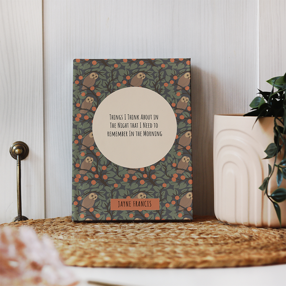 A custom hardback journal with a repeat pattern of a barn owl in trees with fruit. In the middle of the design is a large moon with text, 'Things I think about in the night that I need to remember in the morning'. The journal is propped up against the wall on a grass mat next to a potted plant.