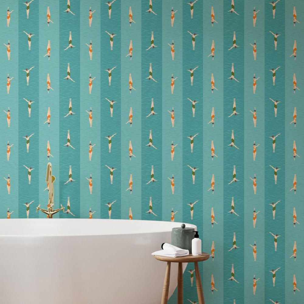 A bathroom with a bath, products on a side table and a repeat striped wallpaper pattern featuring two alternating white women swimmers in orange and green swim costumes